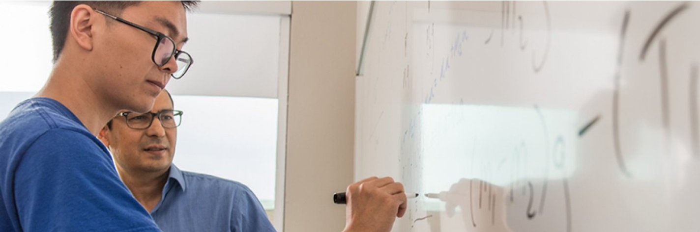 Student writing equations on a board with the professor standing next to him.